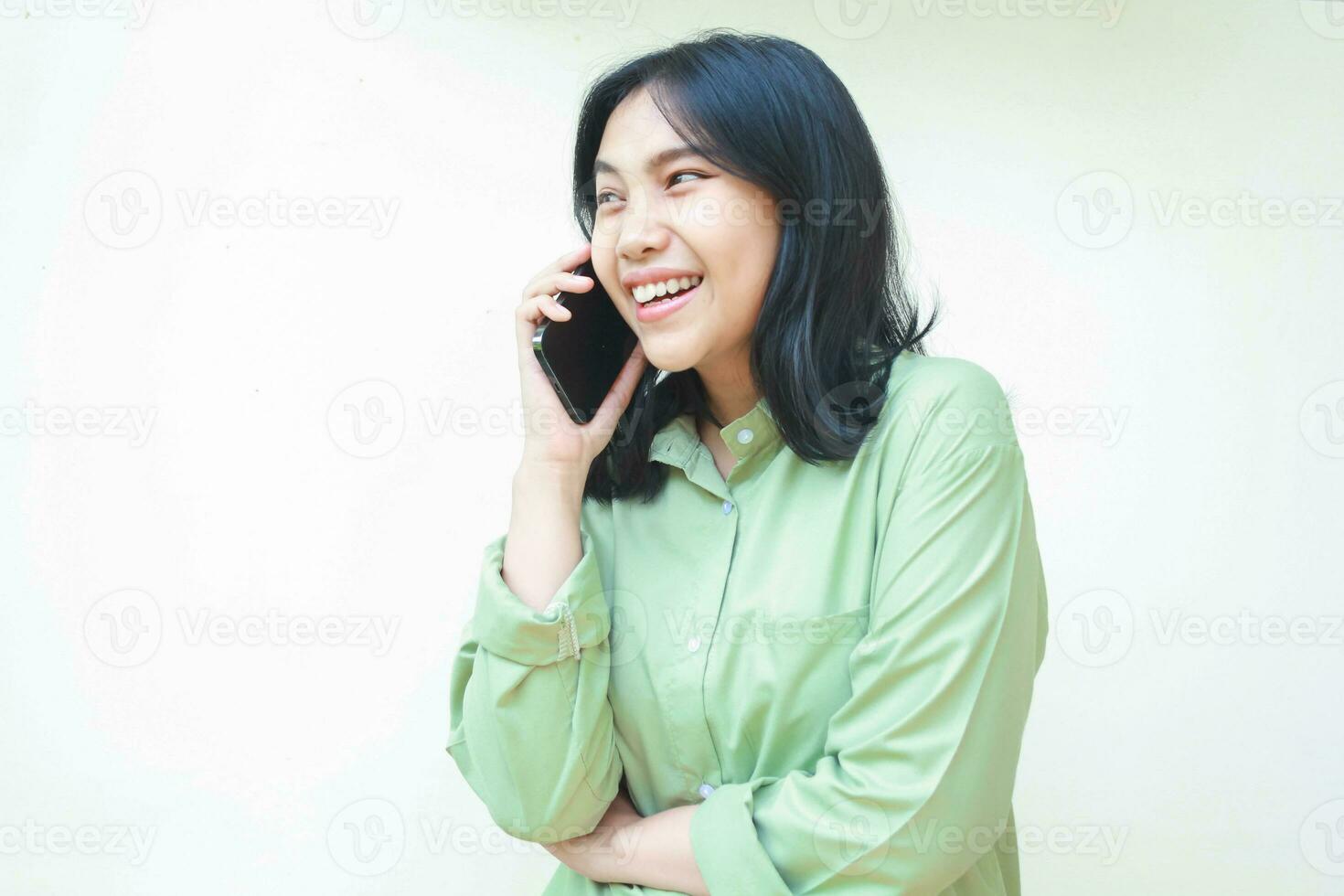 em êxtase bem sucedido ásia mulher vestindo verde grande demais camisa falando em Smartphone rindo do engraçado conversação em pé sobre isolado branco fundo foto