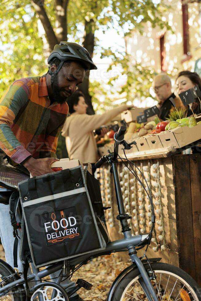 fresco frutas e legumes Entrega. jovem africano masculino correio colocando eco orgânico produzir dentro térmico mochila enquanto colheita acima ordem a partir de agricultores mercado, entregando saudável local Comida para clientes foto