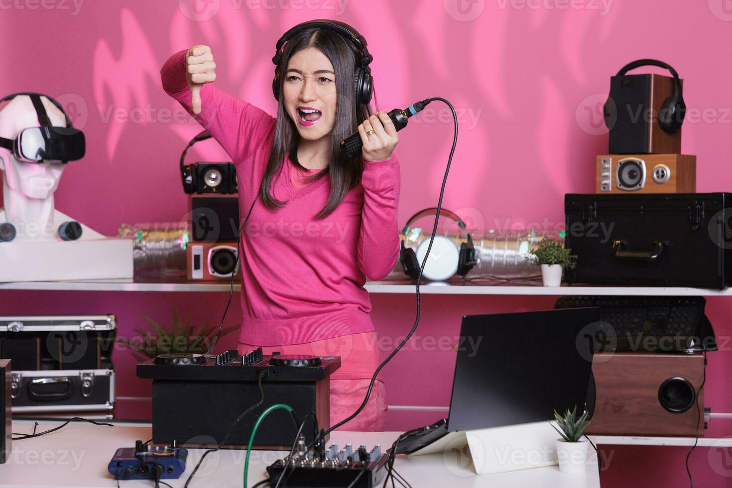 infeliz artista segurando microfone enquanto fazendo desacordo gesto, expressando desacordo dentro estúdio sobre Rosa fundo. dj mulher realizando música com eletrônicos equipamento e audio instrumento foto