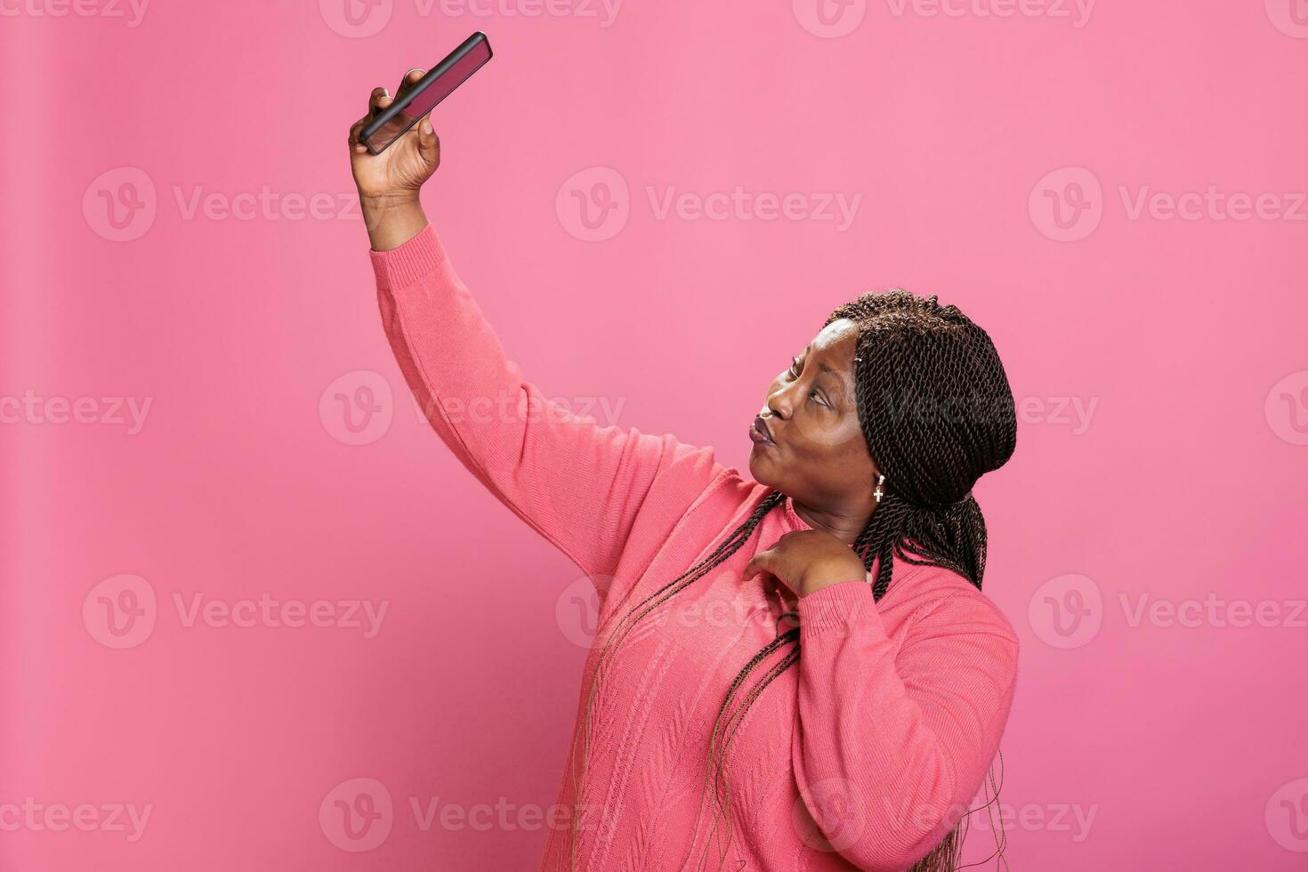 linda africano americano mulher fazendo selfie com Móvel telefone enquanto posando dentro estúdio sobre Rosa fundo. à moda alegre menina levando fotografia desfrutando postagem em social meios de comunicação foto