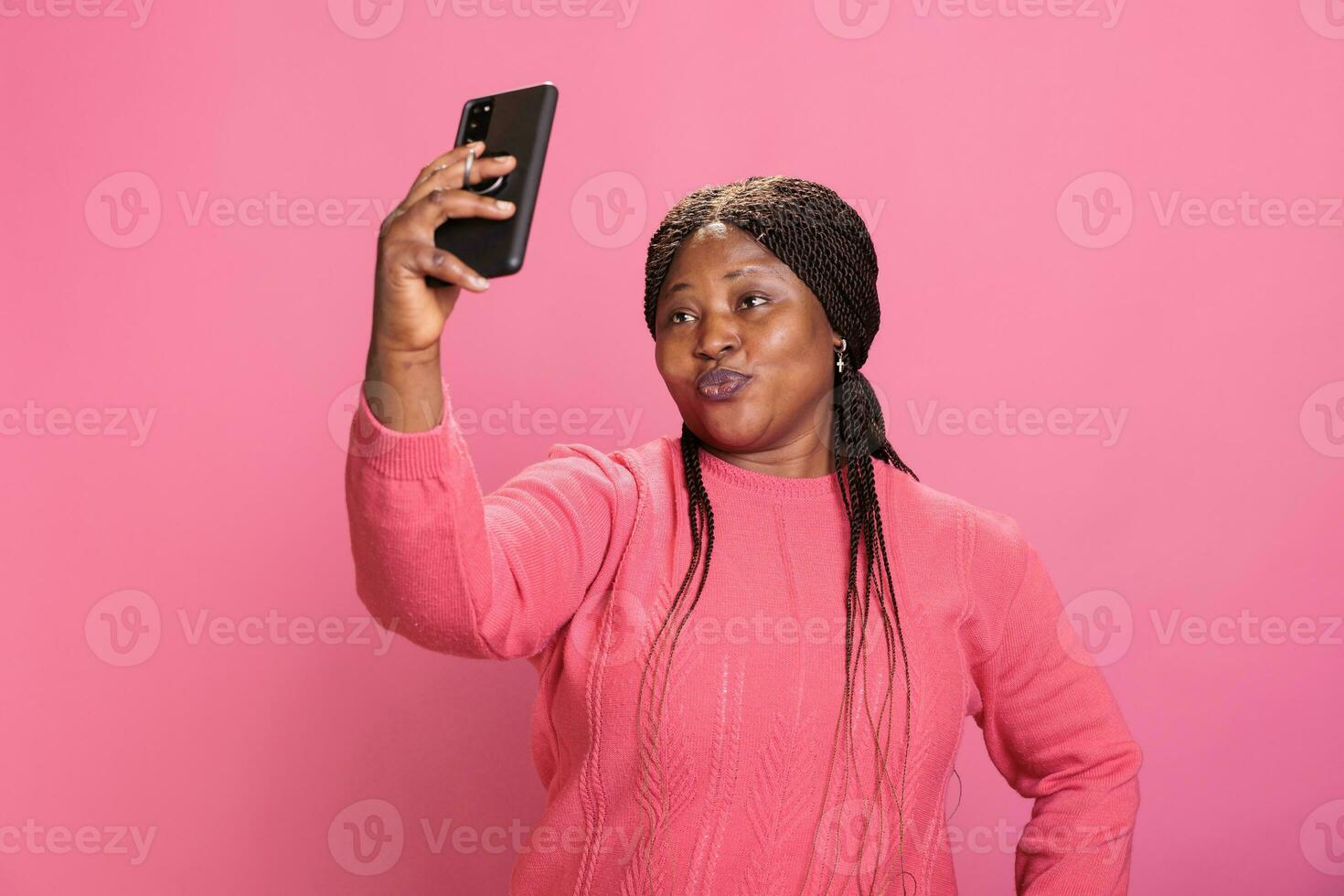 retrato do adorável mulher com à moda Penteado levando selfie usando Smartphone enquanto fazendo engraçado expressão dentro estúdio sobre isolado fundo. alegre atraente pessoa desfrutando fazer fotos