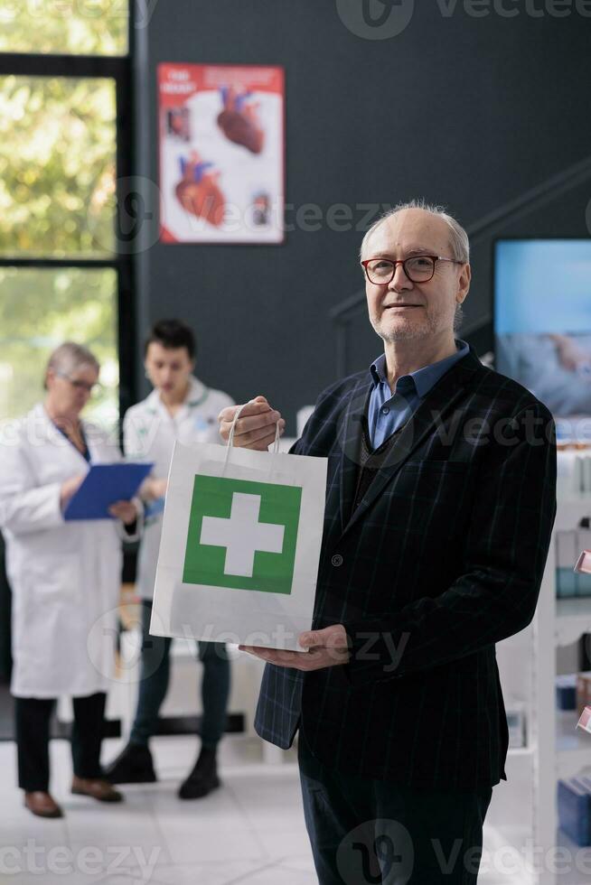 Senior homem segurando compra compras saco dentro farmácia retrato. farmacia idosos sorridente cliente comprando prescrição medicamentos, em pé dentro farmacêutico e olhando às Câmera foto