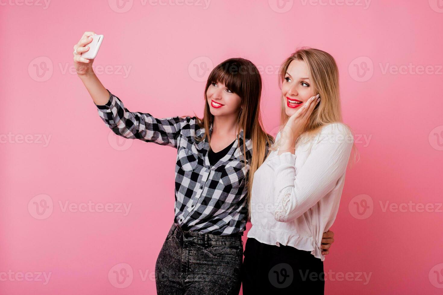 dois bonita meninas fazer auto retrato de Móvel telefone. sorridente, tendo diversão, Rosa fundo. estúdio tomada. foto