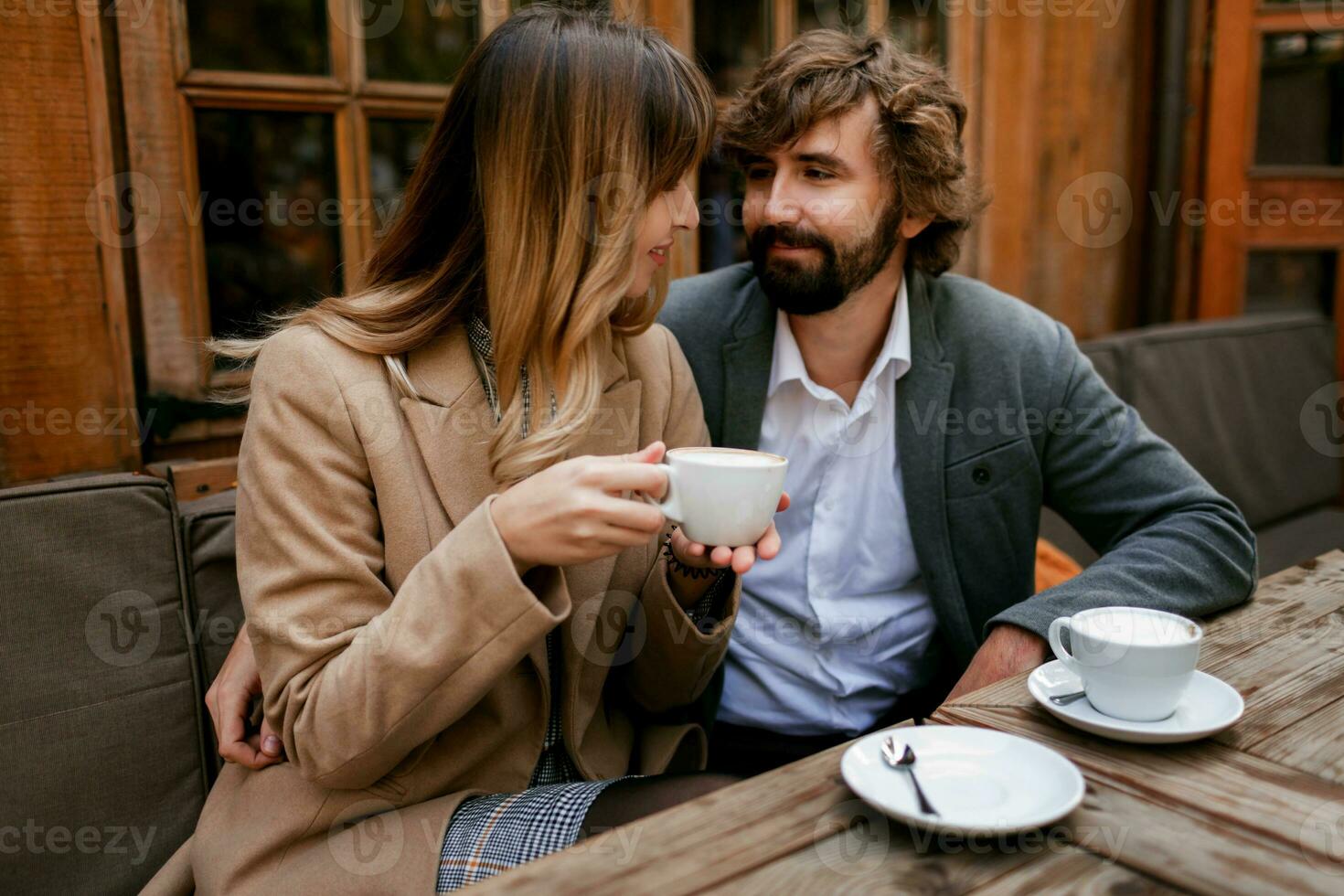 romântico pensativo mulher com grandes ondulado cabelos abraçando dela marido com barba. elegante casal sentado dentro cafeteria com quente cappuccino. foto