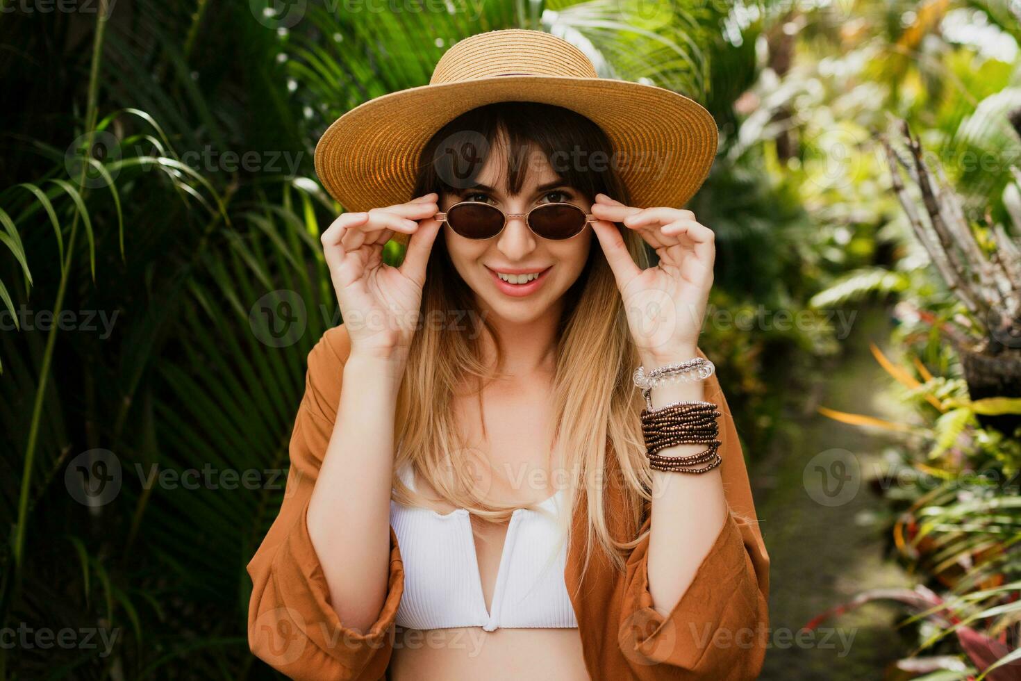fechar acima verão elegante retrato do morena mulher dentro Palha chapéu posando em tropical Palma folhas fundo dentro bali. vestindo à moda boêmio acessórios. foto