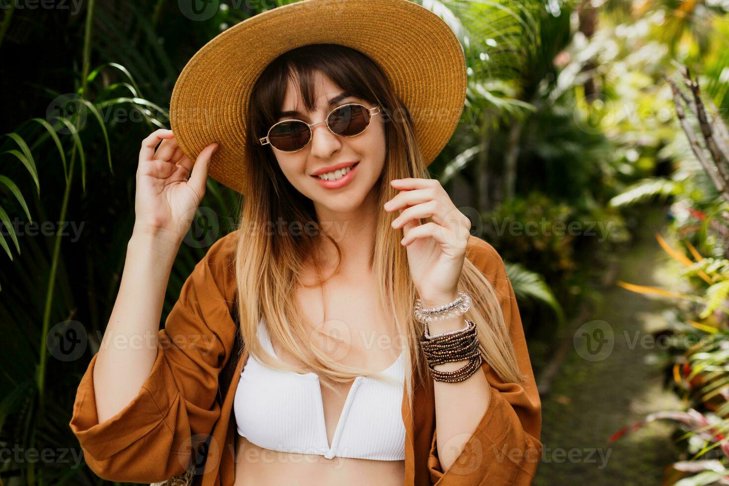 fechar acima verão elegante retrato do morena mulher dentro Palha chapéu posando em tropical Palma folhas fundo dentro bali. vestindo à moda boêmio acessórios. foto