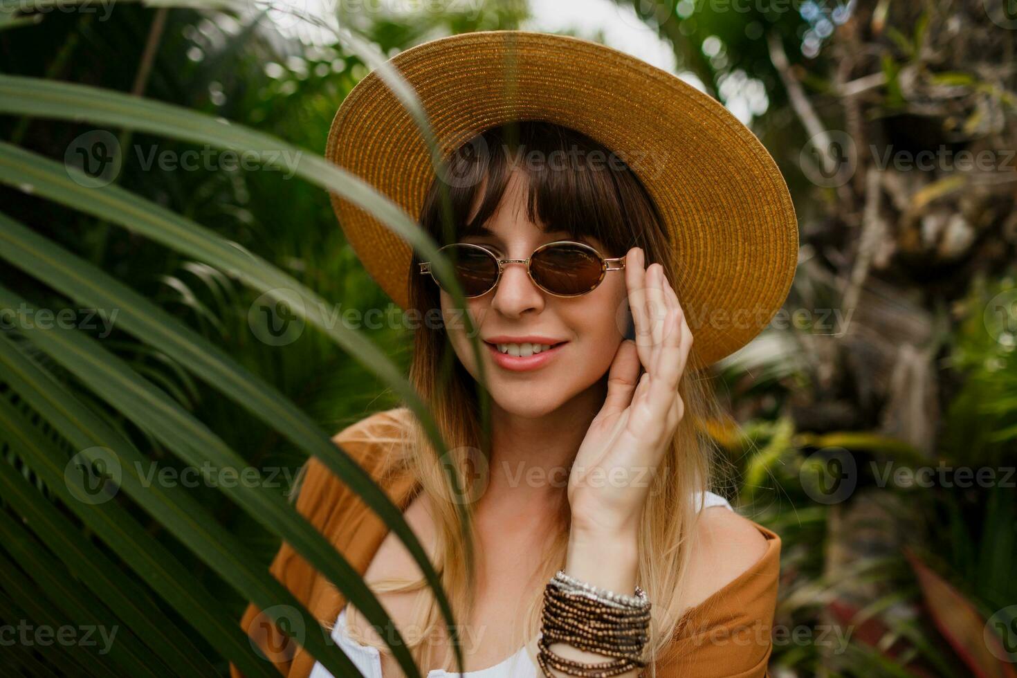 estilo de vida retrato do morena mulher dentro Palha chapéu posando em tropical Palma folhas fundo dentro bali. vestindo à moda boêmio acessórios. foto