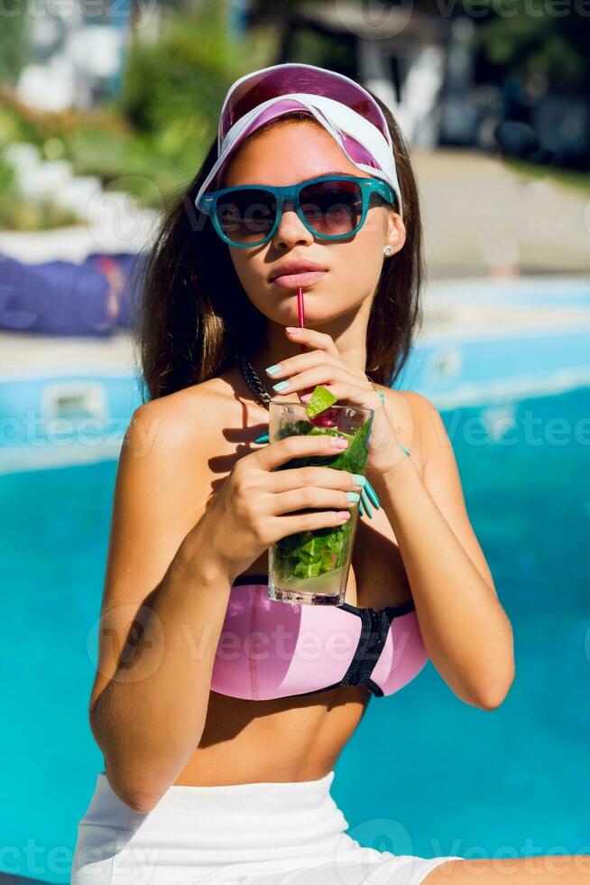 estilo de vida retrato do bronzeada lindo mulher dentro Rosa bikini e oculos de sol sentado perto natação piscina com fresco coquetel. surpreendente sensual menina com perfeito fino esporte corpo posando ar livre. foto