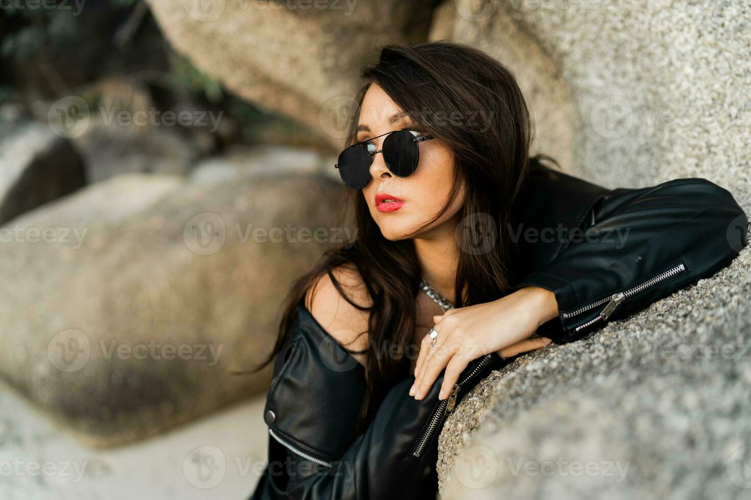 fechar op retrato do confiante mulher com vermelho lábios dentro na moda Preto couro Jaqueta e sensual Preto vestir posando em rochoso fundo. foto