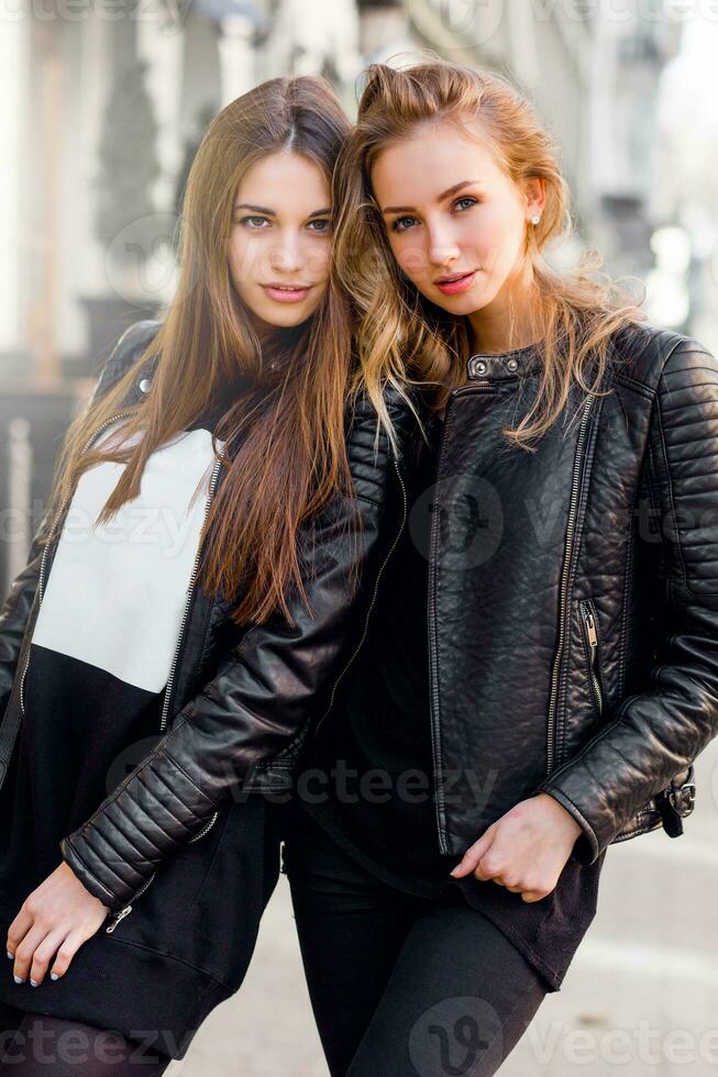 dois à moda bonita meninas posando em a rua dentro ensolarado dia. vestindo na moda urbano equipamento , couro Jaqueta e chuteiras salto. foto