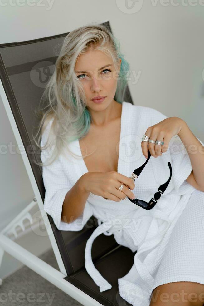 lindo loiro mulher dentro branco roupão arrepiante em área coberta cadeira dentro luxo hotel. perfeito pele, lindo olhos. foto