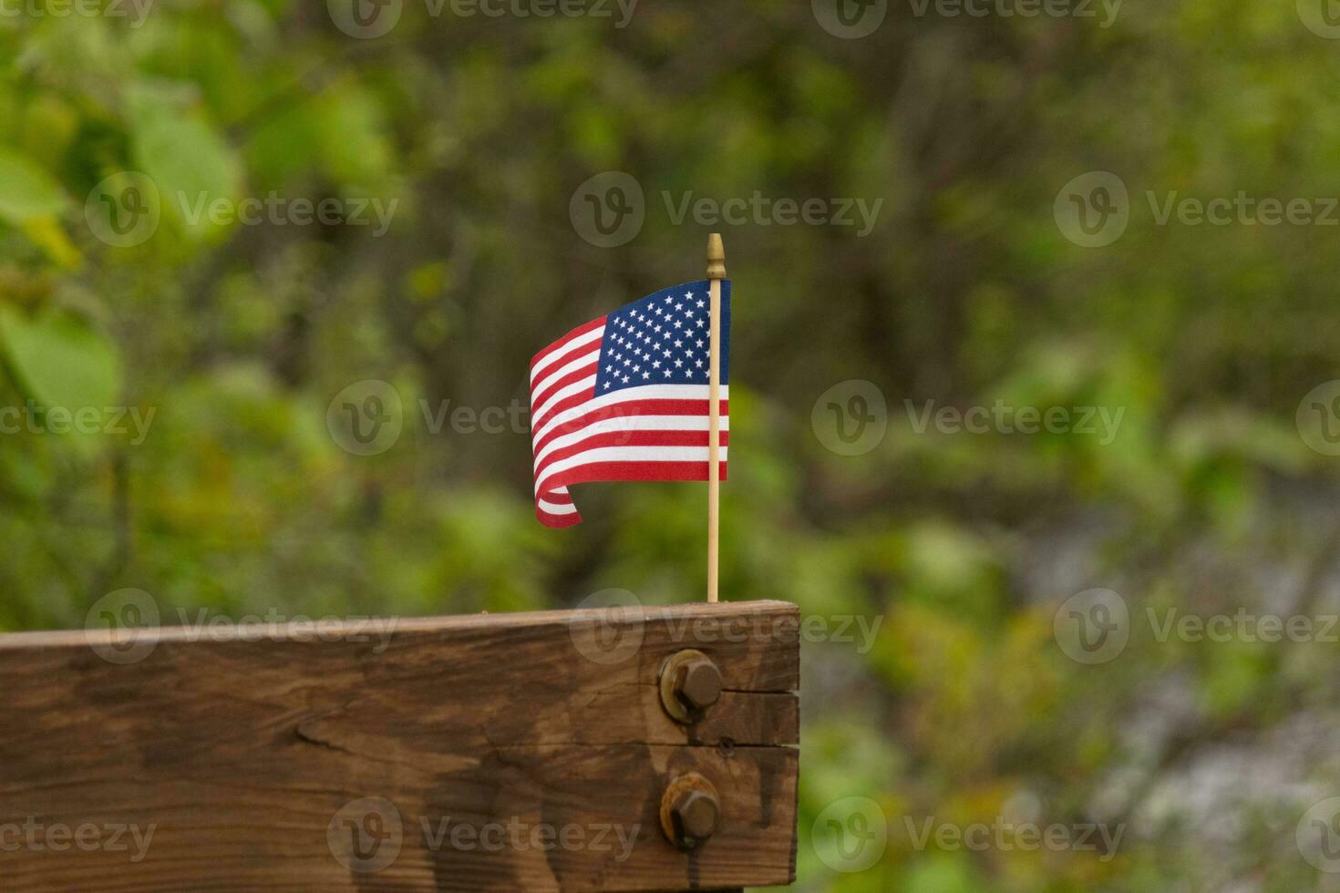 isto é a imagem do uma pequeno americano bandeira fixado para uma de madeira feixe. isto patriótico exibição parece perguntar colorida com a vermelho, branco, e azul. a símbolo do americano é suavemente fluindo dentro a brisa. foto
