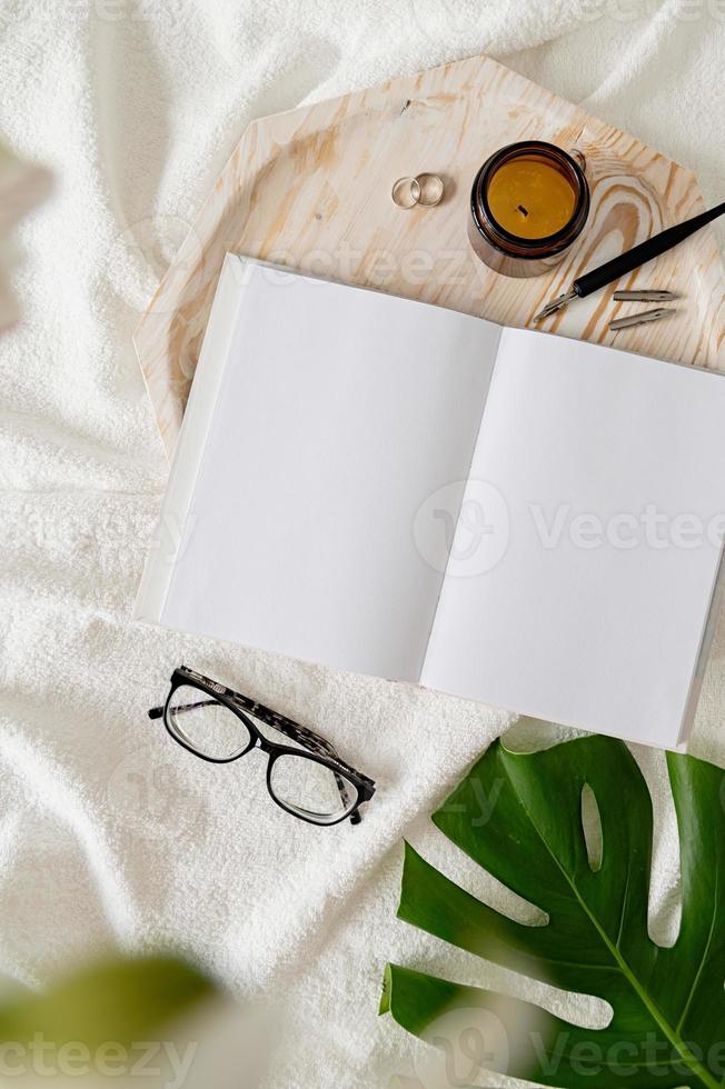 livro aberto, óculos, vela e flores vista de cima na cama branca foto