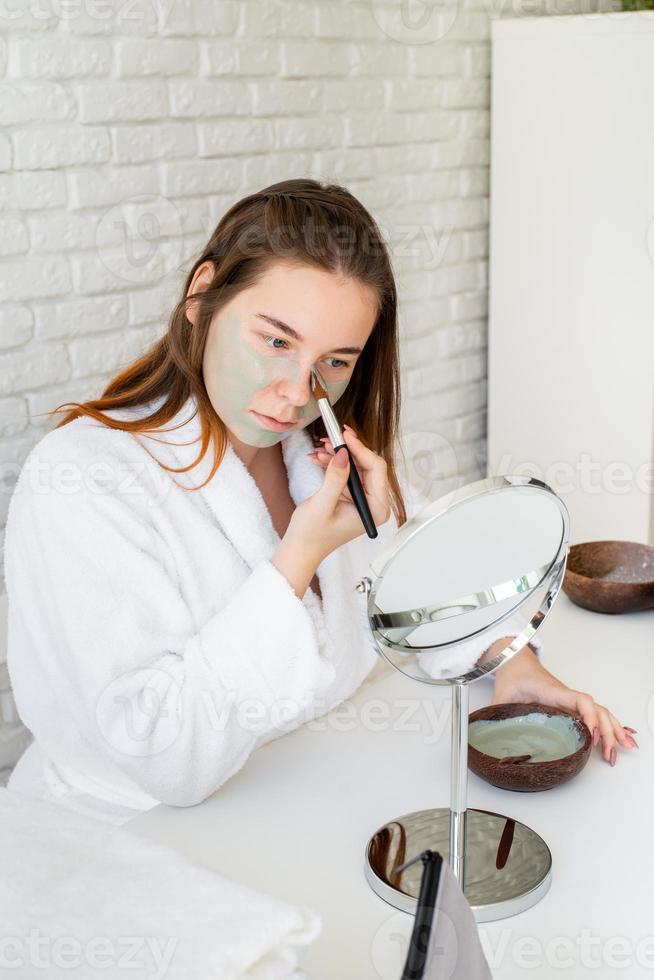 mulher vestindo roupão de banho aplicando máscara facial de argila, olhando para o espelho foto