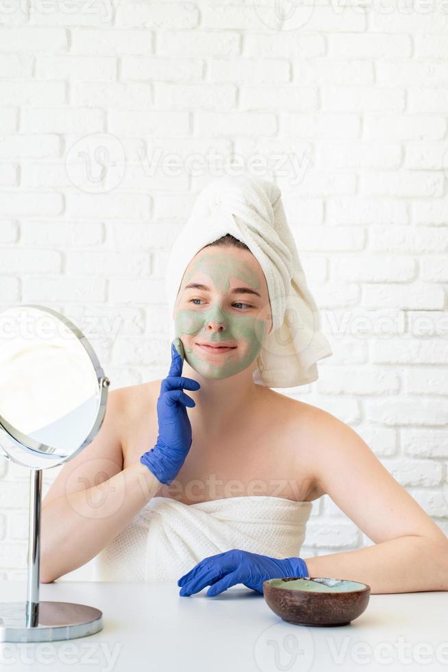 mulher usando luvas, aplicando máscara facial de argila, olhando para o espelho foto