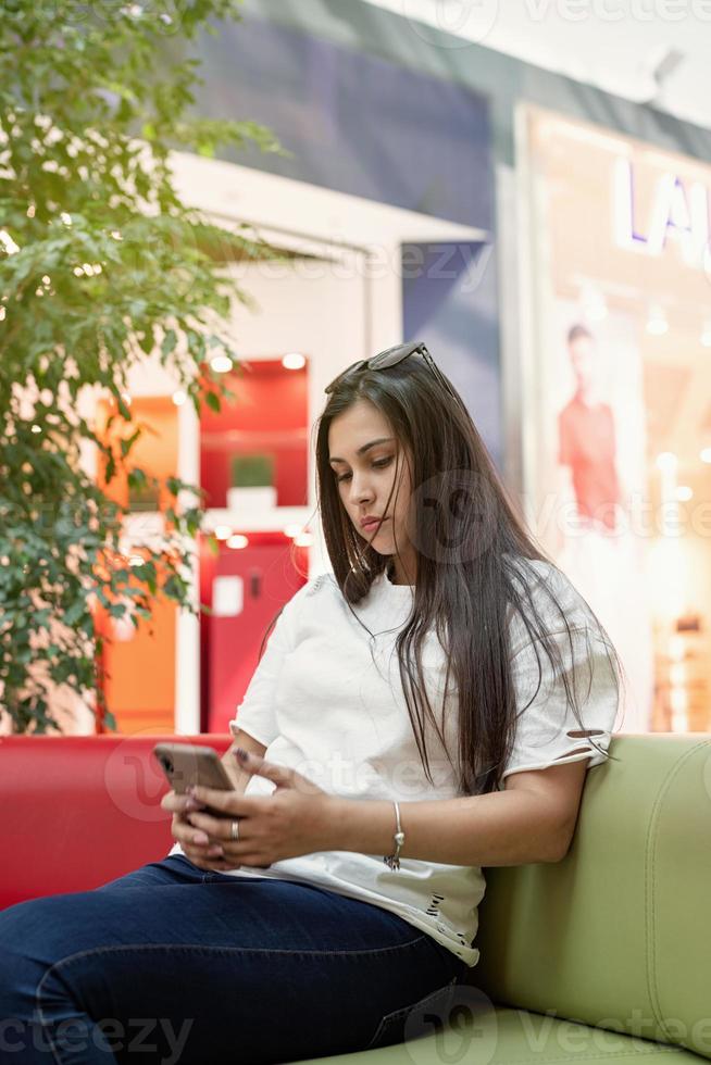 mulher sentada no sofá de um shopping, olhando para o telefone foto