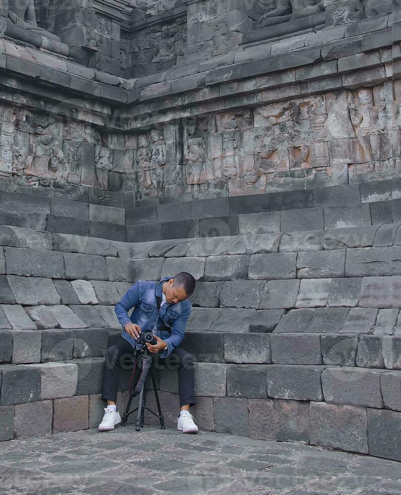 indonésio masculino fotógrafo vestindo uma azul levis Jaqueta usando uma Câmera e tripé sentado dentro a fundo do a borobudur têmpora relevos. foto