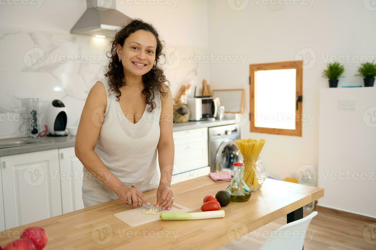 lindo mulher dona de casa sorrisos olhando às Câmera, cortar alho-poró e alho em corte borda dentro minimalista casa cozinha foto