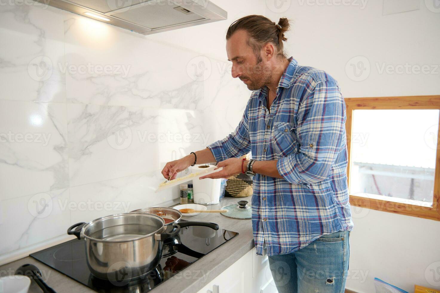 bonito jovem homem colocando picado alho a partir de corte borda para dentro uma panela com ebulição tomate molho, cozinhando às casa foto