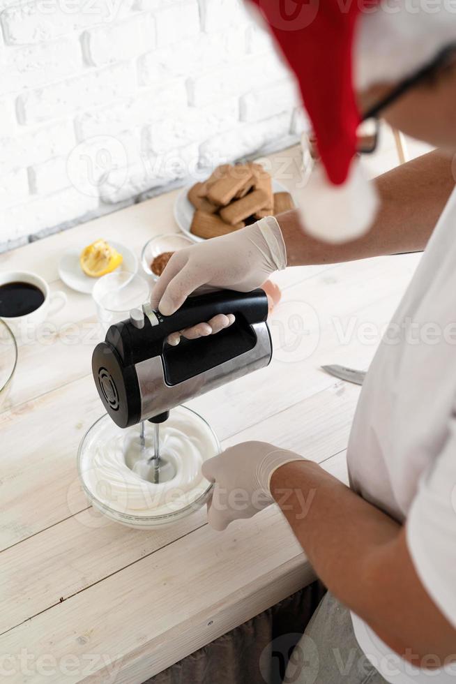 homem com chapéu de Papai Noel batendo claras de ovo com a batedeira na cozinha foto