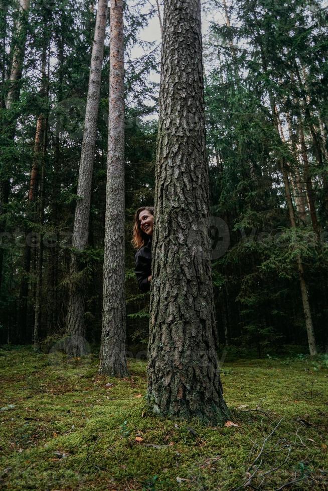 uma jovem em uma floresta de coníferas olha por trás de uma árvore foto