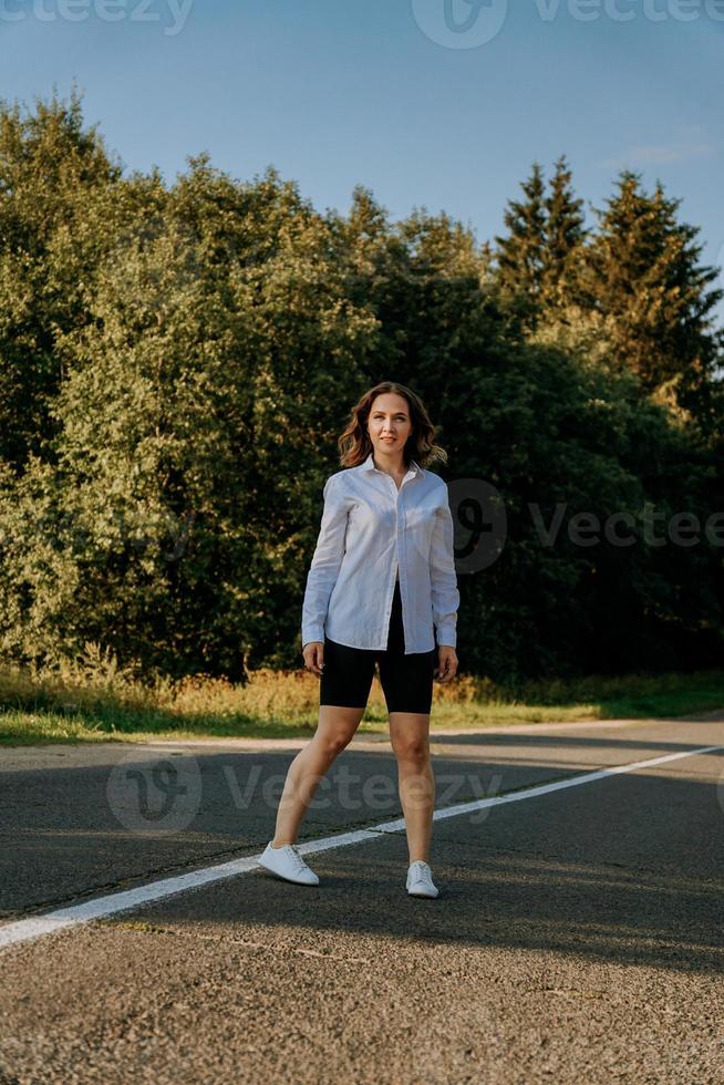 mulher em uma camisa branca caminha ao longo da estrada entre a floresta foto