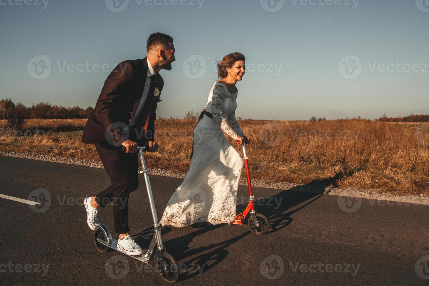 casal de noivos sorridente andando de scooters ao longo da estrada ao pôr do sol foto