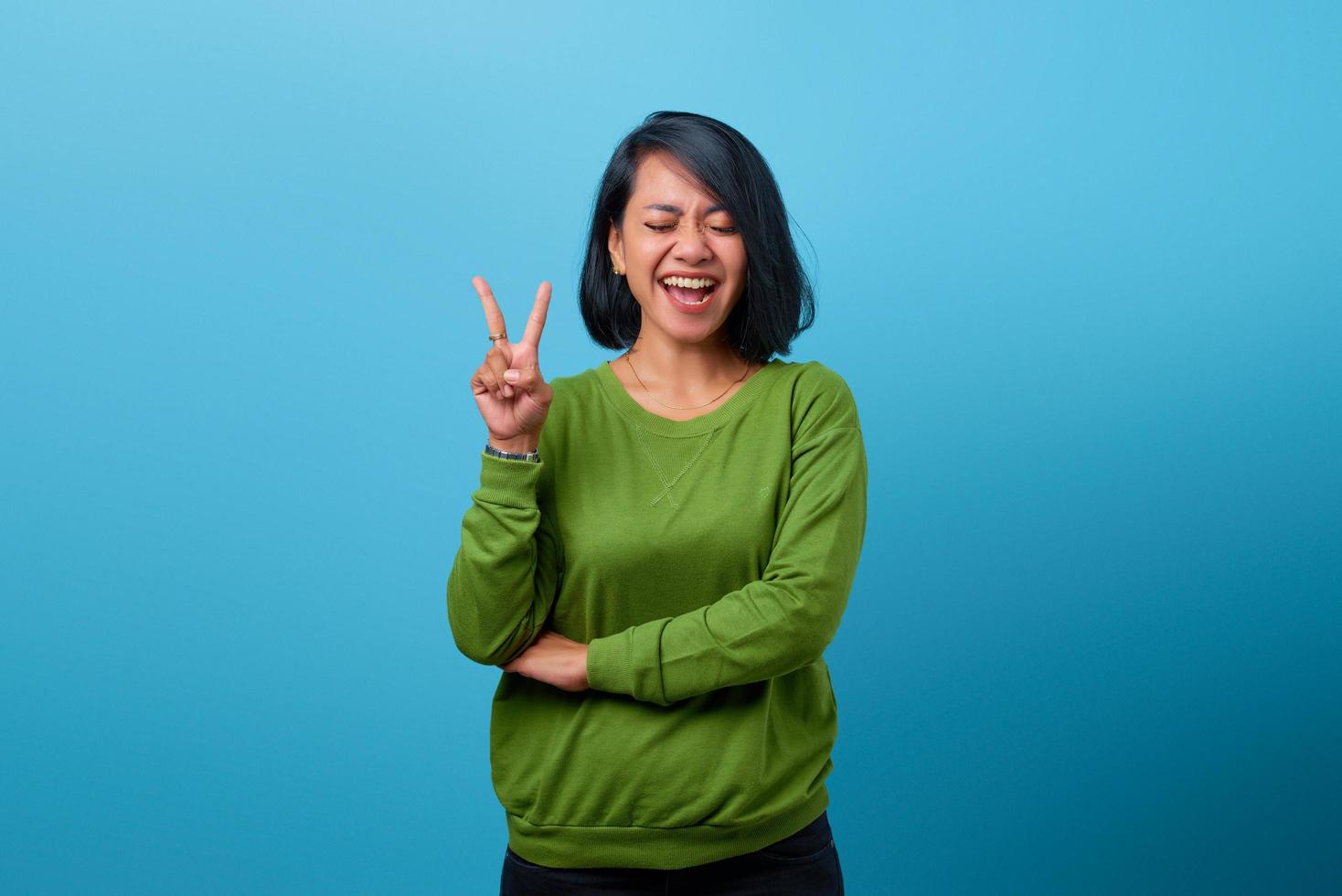 Mulher asiática atraente sorrindo e fazendo sinal de vitória sobre fundo azul foto