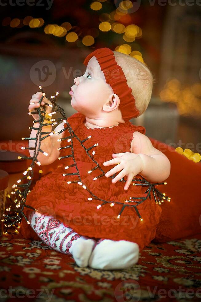 pequeno menina dentro vermelho vestir contra fundo do Natal árvore detém Natal festão dentro dela mãos. bebê 6 mês velho comemora Natal. foto