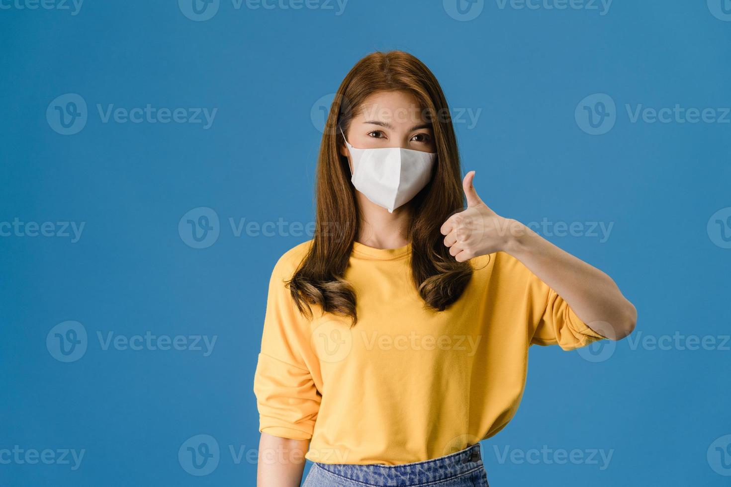 jovem asiática usa máscara facial aparecendo o polegar sobre fundo azul. foto