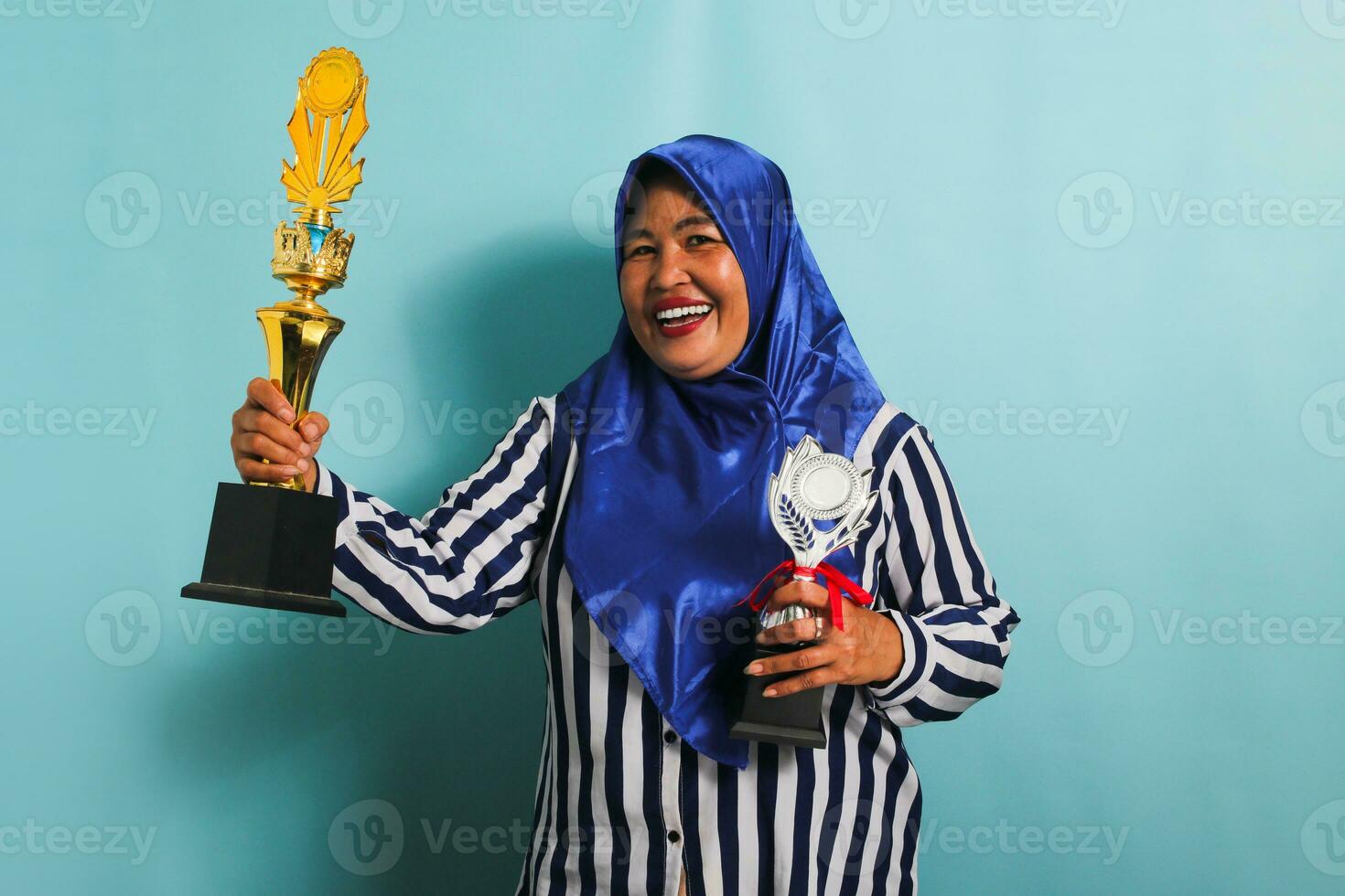 a animado meia idade ásia empresária dentro uma azul hijab e uma listrado camisa é segurando uma ouro e prata troféu, a comemorar dela sucesso e conquista. ela é isolado em uma azul fundo foto