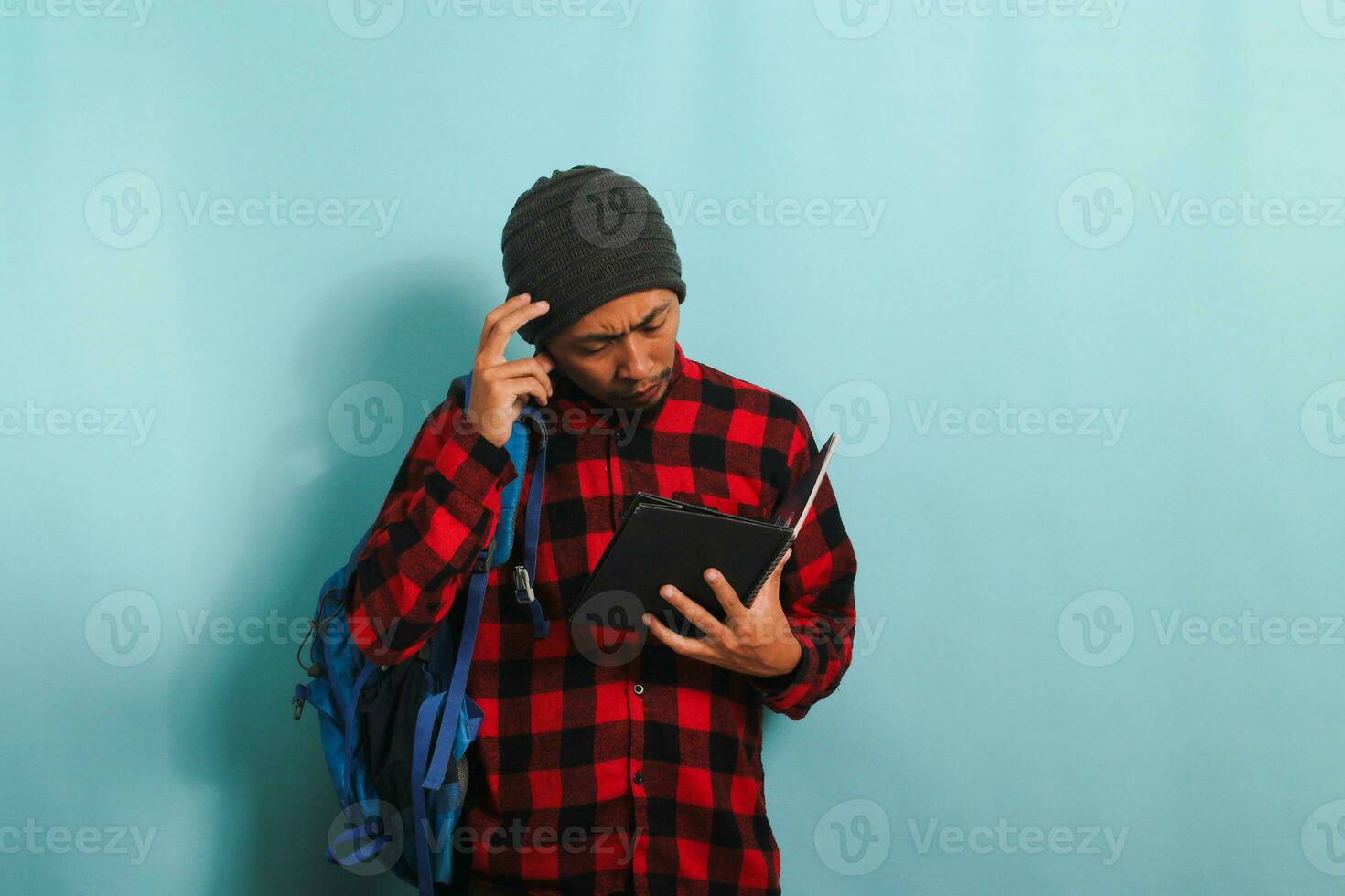 uma pensativo jovem ásia aluna com uma gorro chapéu e uma vermelho xadrez flanela camisa, vestindo uma mochila, é profundo dentro pensamento, coçar dele cabeça olhando para responda enquanto em pé contra uma azul fundo foto