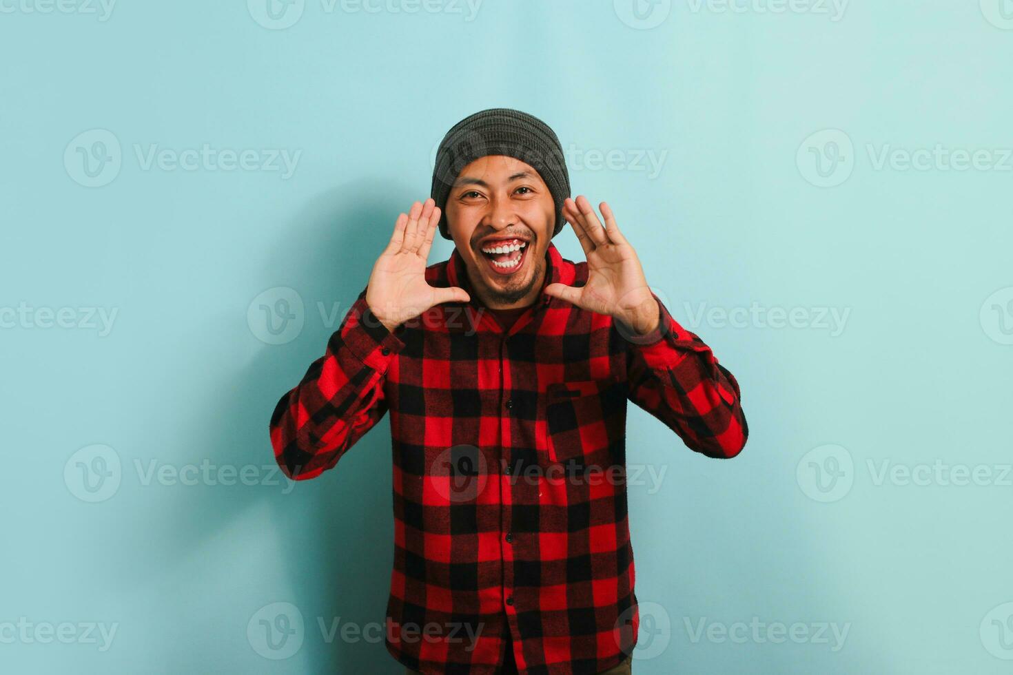 animado jovem ásia homem com uma gorro chapéu e vermelho xadrez flanela camisa gritando e segurando dele Palma perto dele aberto boca, dizendo Boa notícias, isolado em uma azul fundo foto