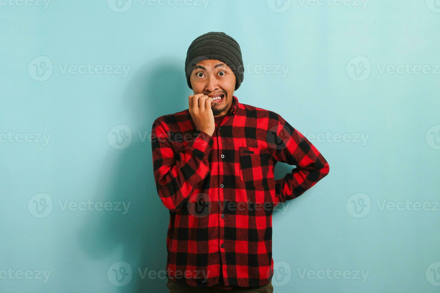 jovem ásia homem com uma gorro chapéu e vermelho xadrez flanela camisa mordidas dele unhas dentro perplexidade, olhando nervosamente às a Câmera, isolado em uma azul fundo foto