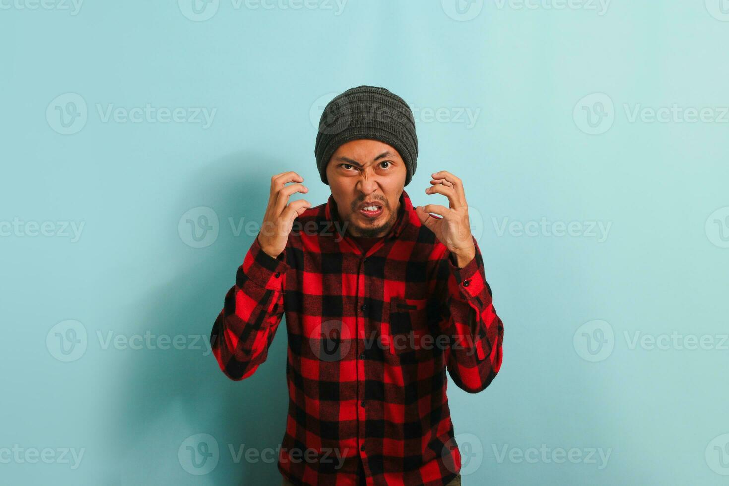 a Bravo jovem ásia homem com uma gorro chapéu e uma vermelho xadrez flanela camisa detém dele mãos dentro uma furioso gesto, gritando às a Câmera. ele é isolado em uma azul fundo foto