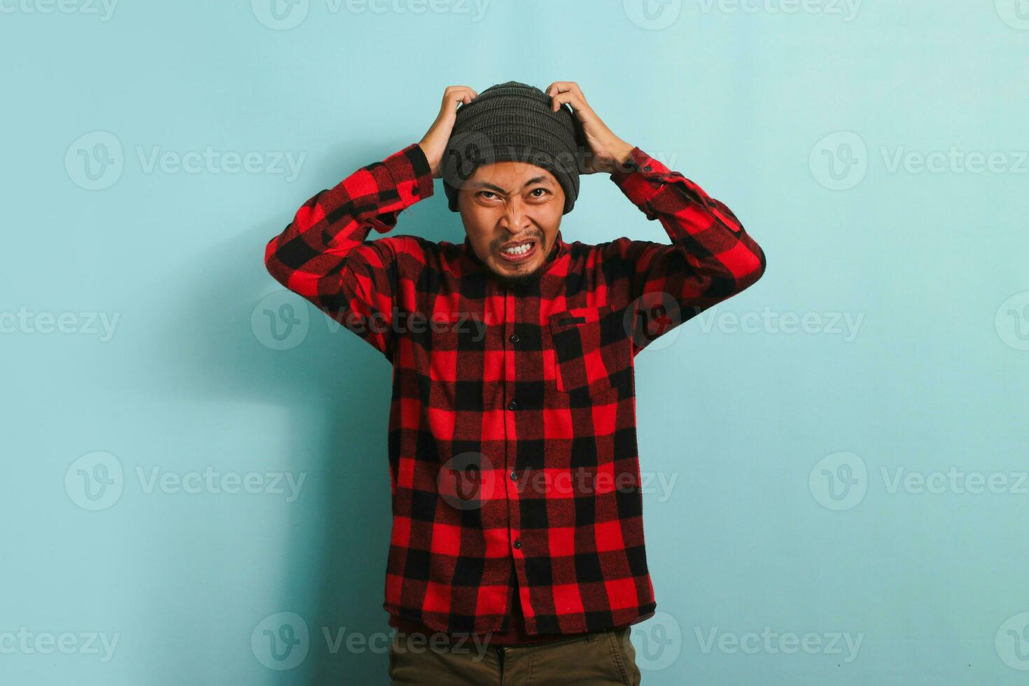 a Bravo jovem ásia homem com uma gorro chapéu e uma vermelho xadrez flanela camisa agarra dele cabeça, sentindo-me descontente, chateado, e desapontado. ele é isolado em uma azul fundo foto