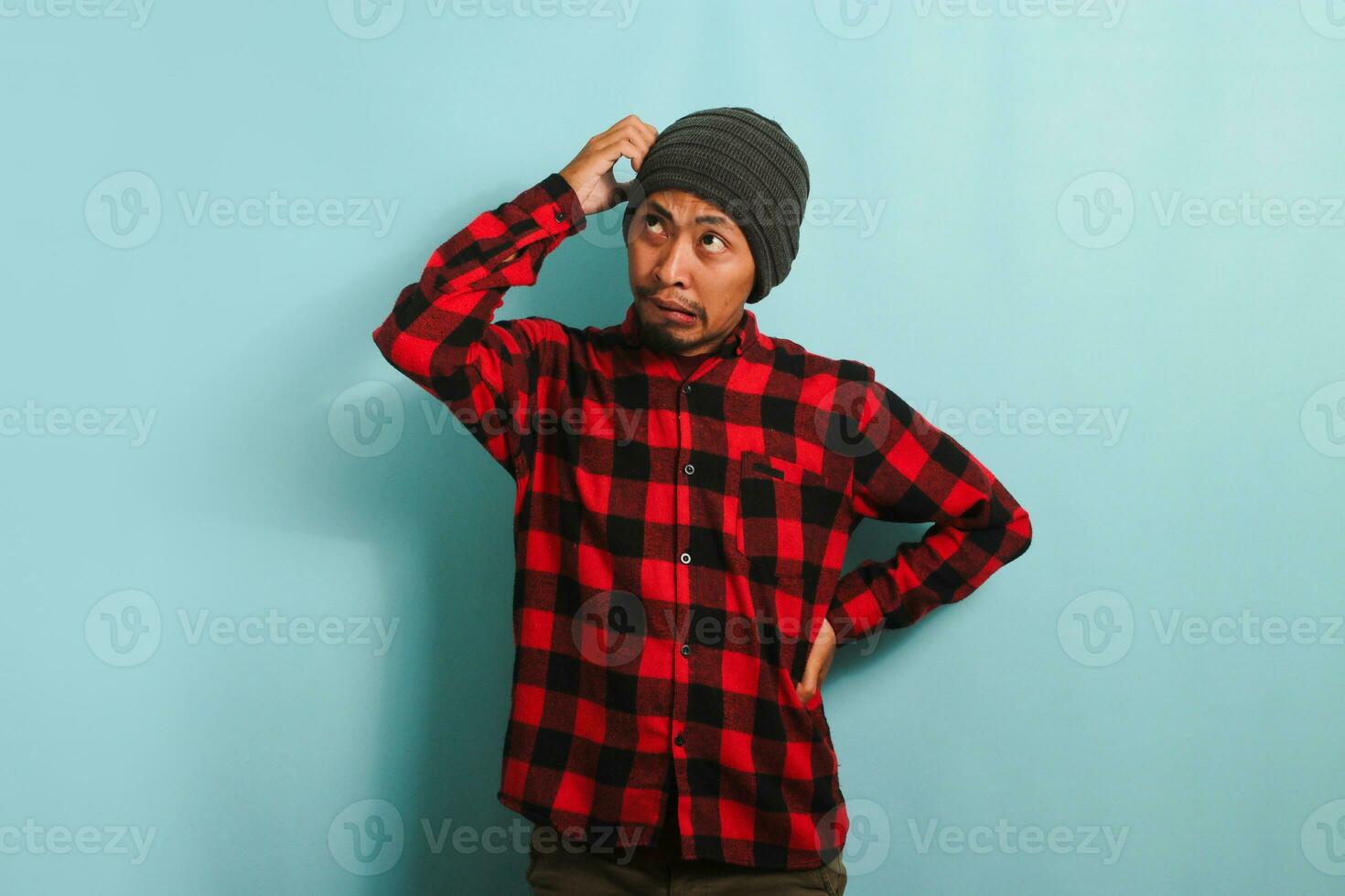 uma confuso jovem ásia homem com uma gorro chapéu e uma vermelho xadrez flanela camisa coçar dele cabeça e olhando acima às a cópia de espaço enquanto em pé contra uma azul fundo foto