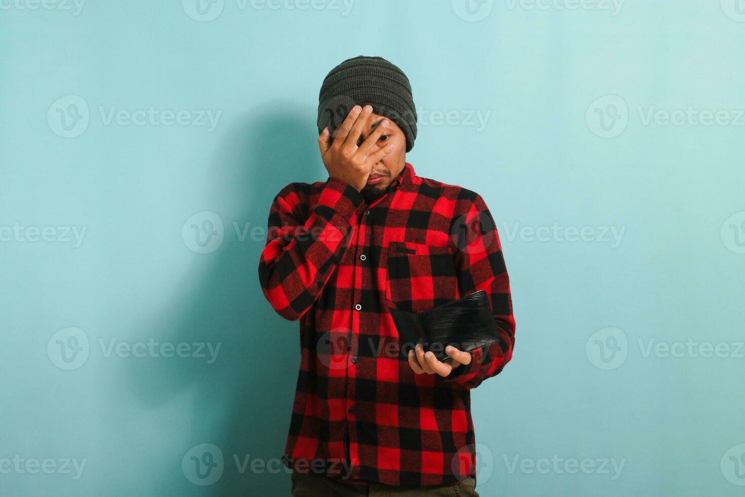 engraçado jovem ásia homem com gorro chapéu e vermelho xadrez flanela camisa cobertura dele face com dele mão e espreitar entre dele dedos enquanto olhando às dele esvaziar carteira, isolado em uma azul fundo foto