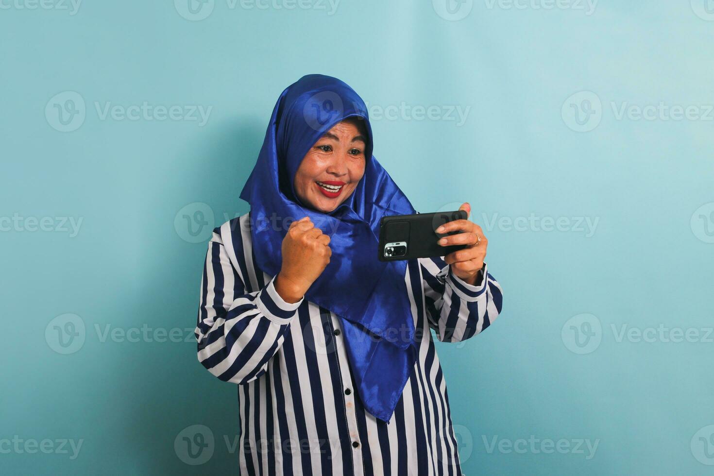 animado meia idade ásia mulher dentro uma azul hijab e uma listrado camisa, jogando uma jogos em Móvel telefone, isolado em uma azul fundo. foto