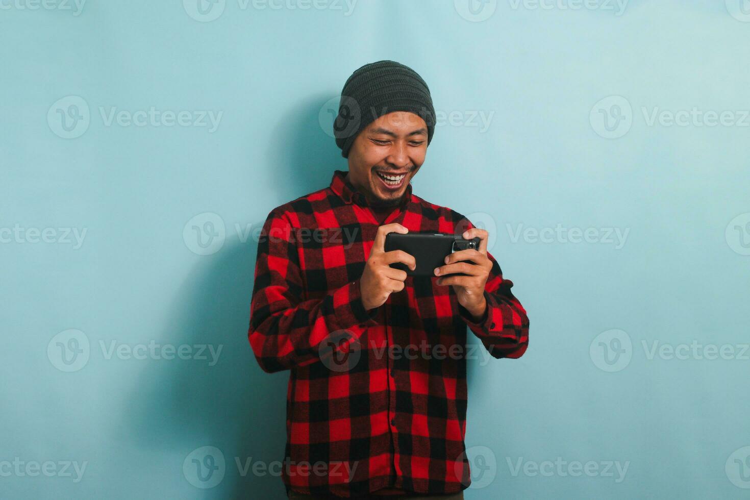 animado jovem ásia homem com uma gorro chapéu e uma vermelho xadrez flanela camisa é jogando a conectados jogos em dele Smartphone, isolado em uma azul fundo foto