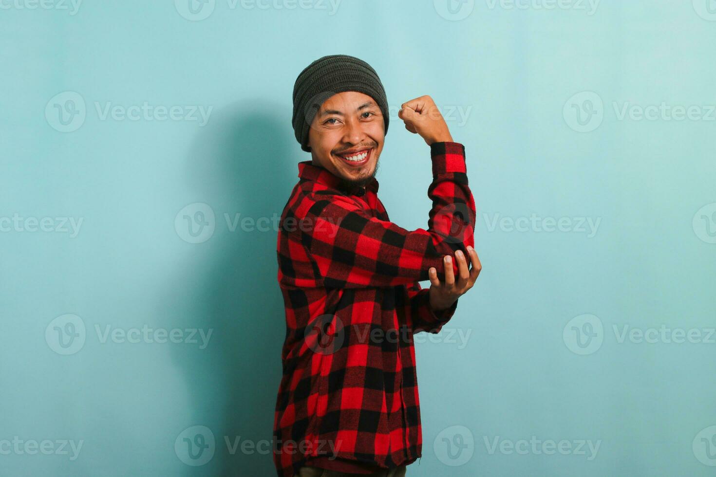 animado jovem ásia homem com gorro chapéu e vermelho xadrez flanela camisa fazer uma Forte gesto, mostrando força de flexão dele braços e músculos, isolado em uma azul fundo foto