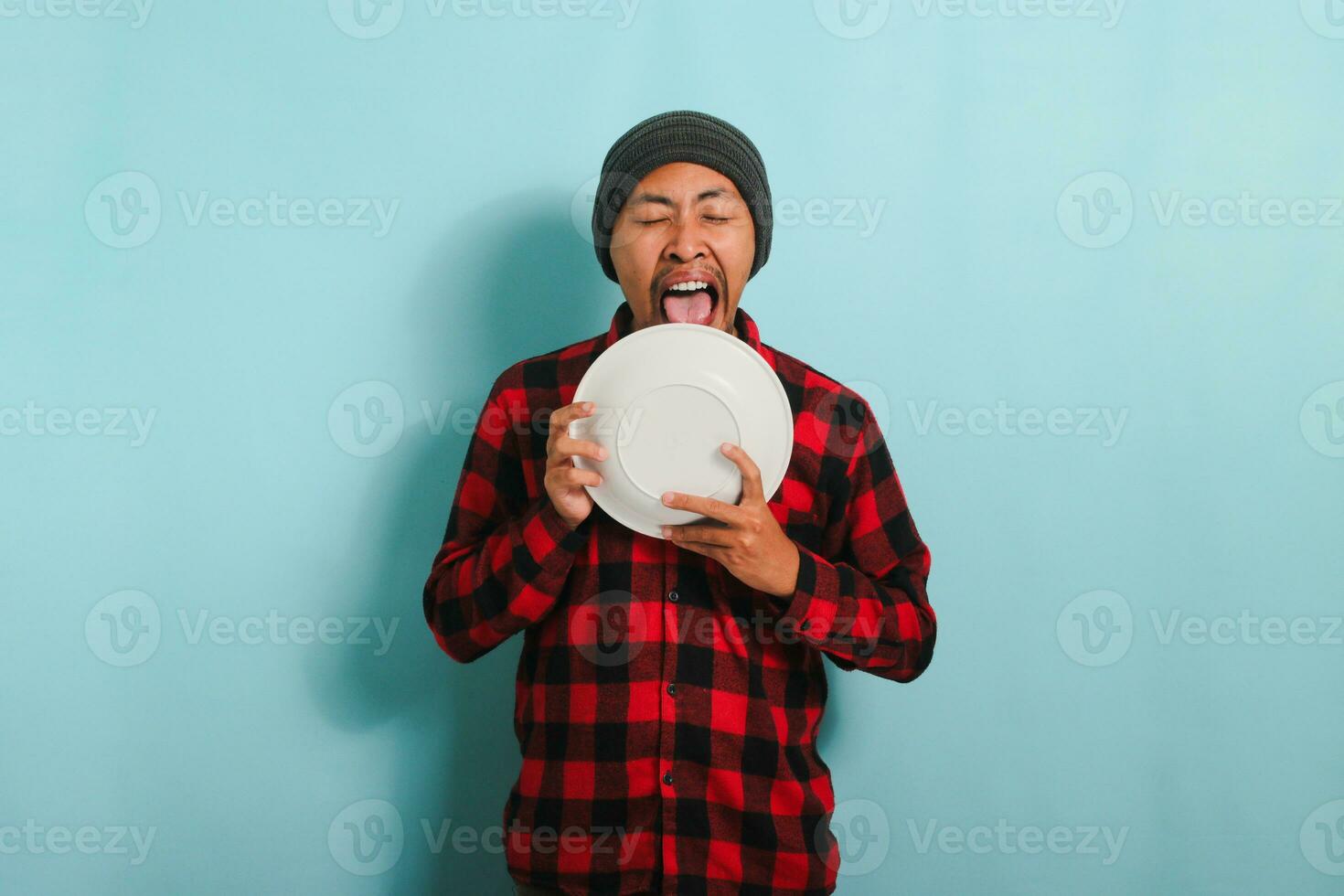 com fome jovem ásia homem com uma gorro chapéu e uma vermelho xadrez flanela camisa é lambendo a placa, indicando este ele é ainda com fome, isolado em uma azul fundo foto