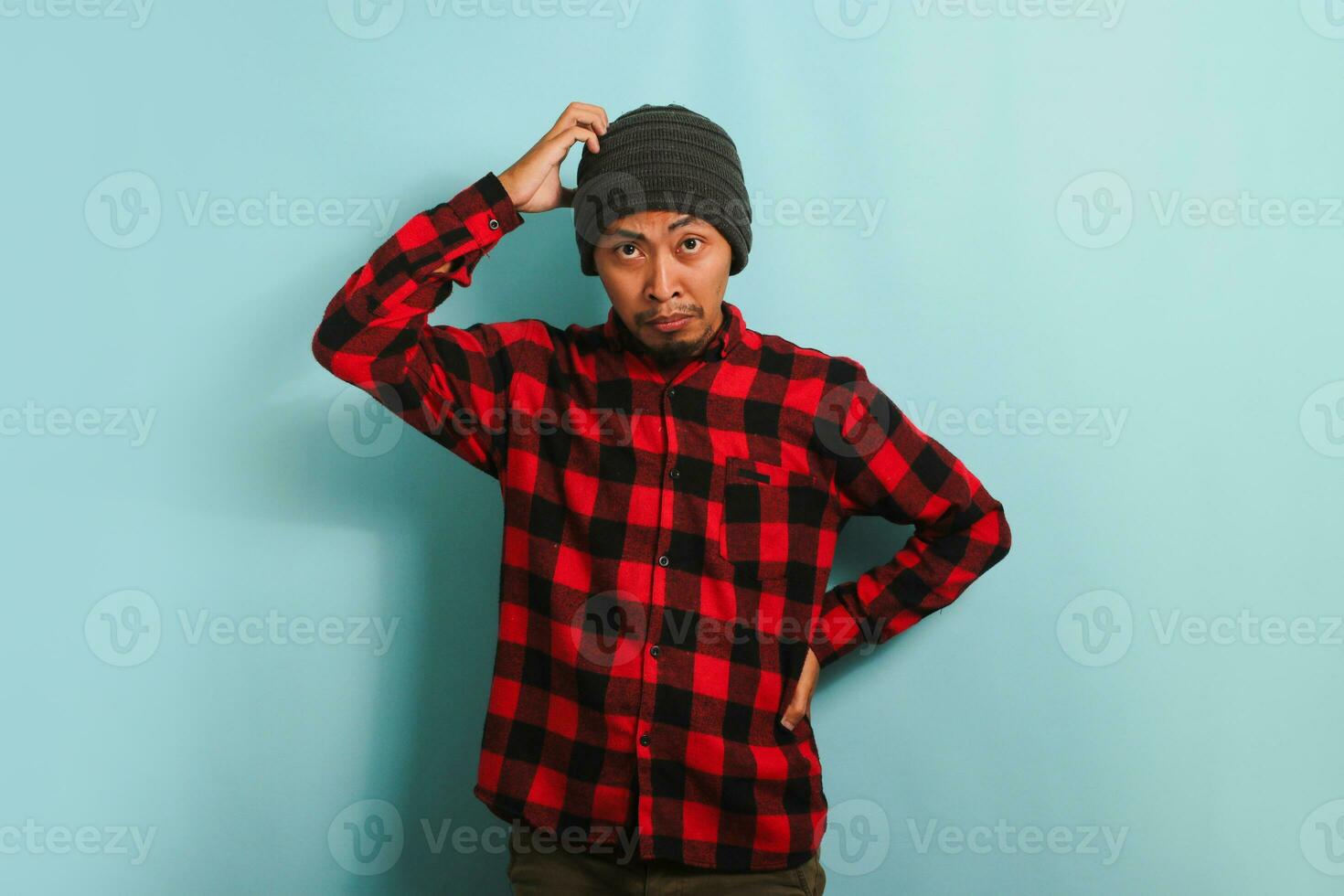 pensativo jovem ásia homem com uma gorro chapéu e vermelho xadrez flanela camisa é coçar dele cabeça, profundo dentro pensamento, tentando para encontrar uma solução, isolado em uma azul fundo foto