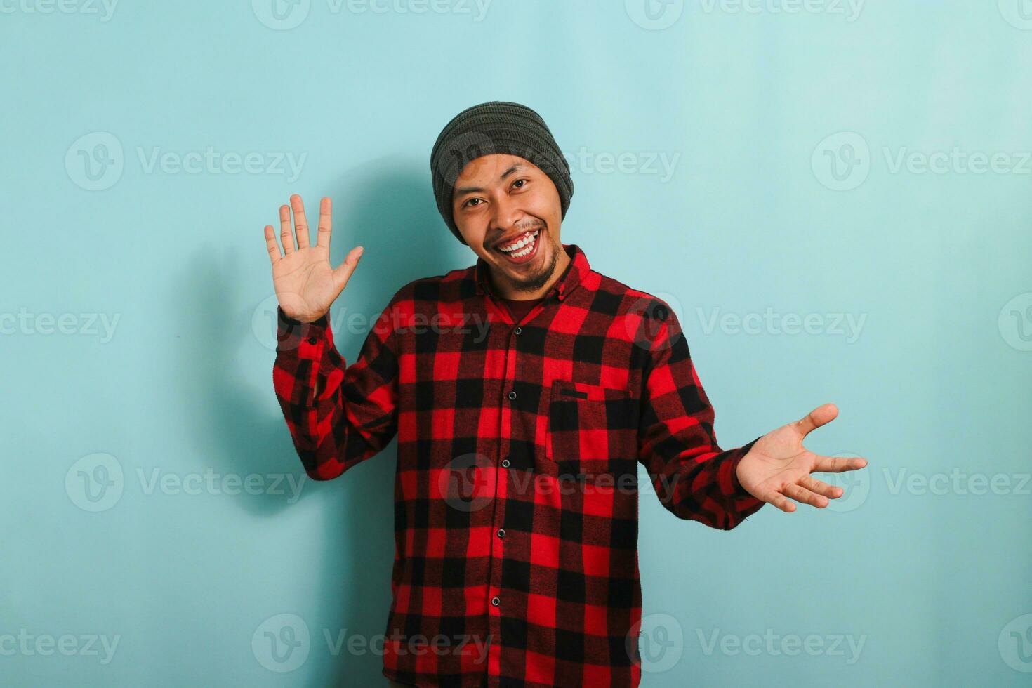 uma sorridente jovem ásia homem com uma gorro chapéu e uma vermelho xadrez flanela camisa puxa dele mãos para a Câmera, convidativo alguém dentro com uma acolhedor gesto enquanto em pé contra uma azul fundo foto