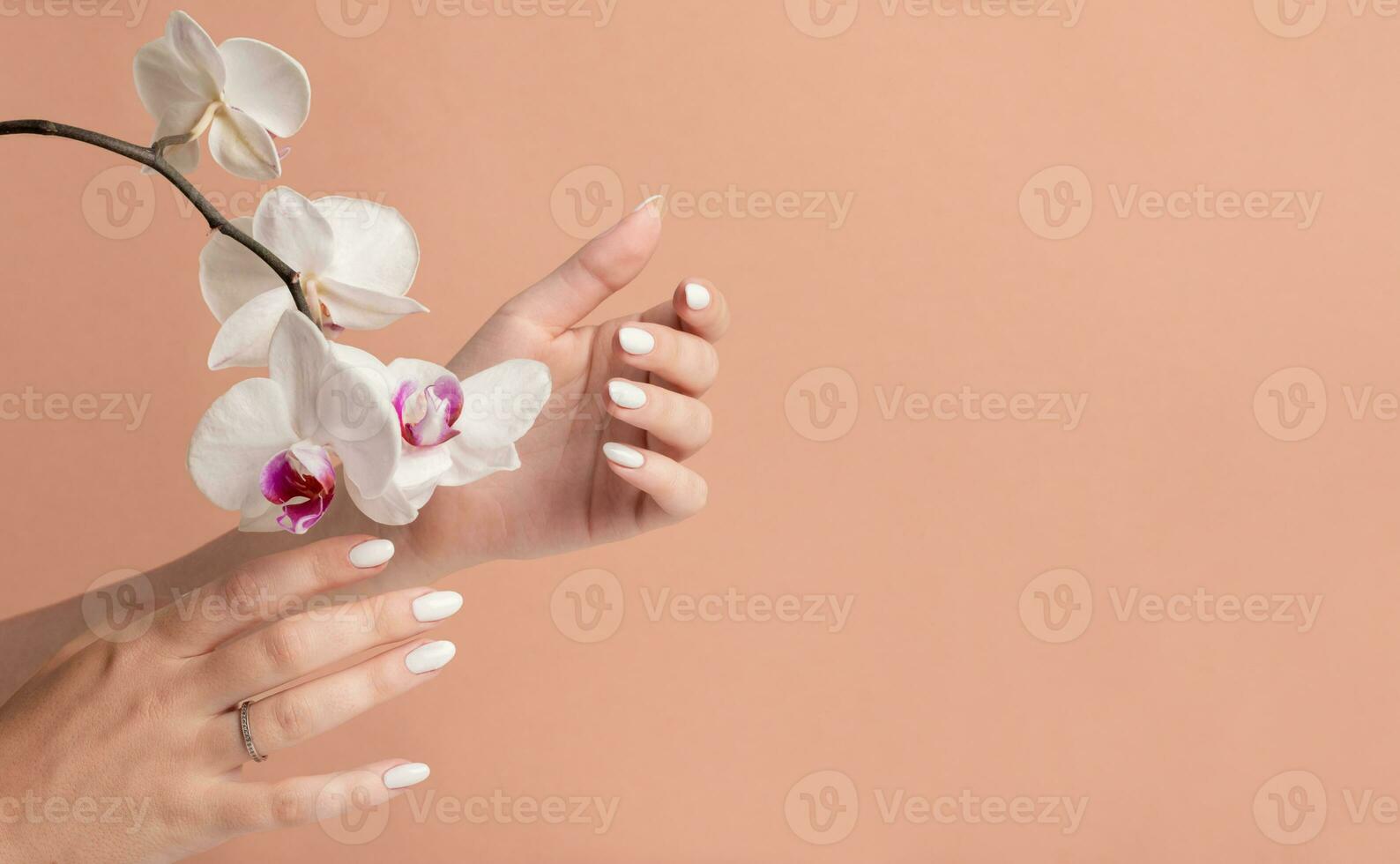 mãos do uma jovem mulher com branco grandes unhas em uma bege fundo com orquídea flores manicure foto