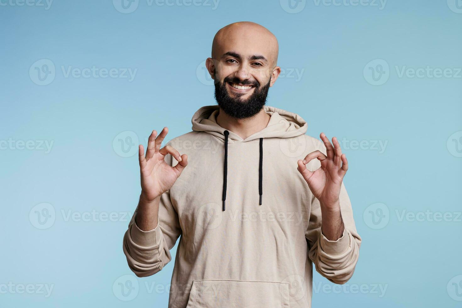 sorridente árabe homem mostrando Está bem gesto com dois mãos e olhando às Câmera. alegre árabe barbudo pessoa posando com OK e acordo placa com dedos estúdio retrato foto