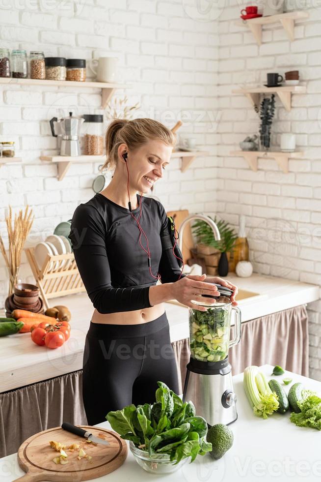 jovem loira sorridente fazendo suco verde na cozinha de casa foto