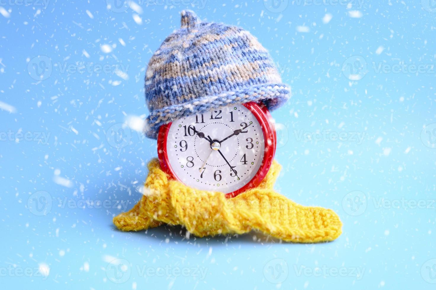 despertador redondo vermelho com chapéu de malha de lã azul e lenço amarelo sobre fundo azul. conceito de inverno. Inverno. aconchegante e aconchegante. tempo analógico 10 10. neve caindo foto