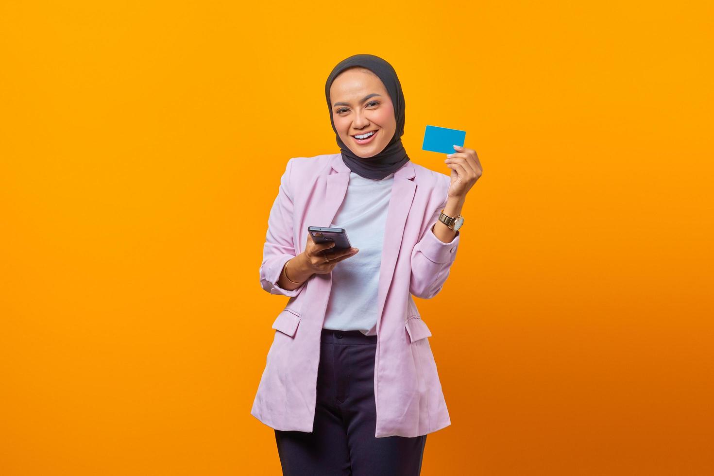 alegre mulher asiática segurando um smartphone e mostrando um cartão em branco foto