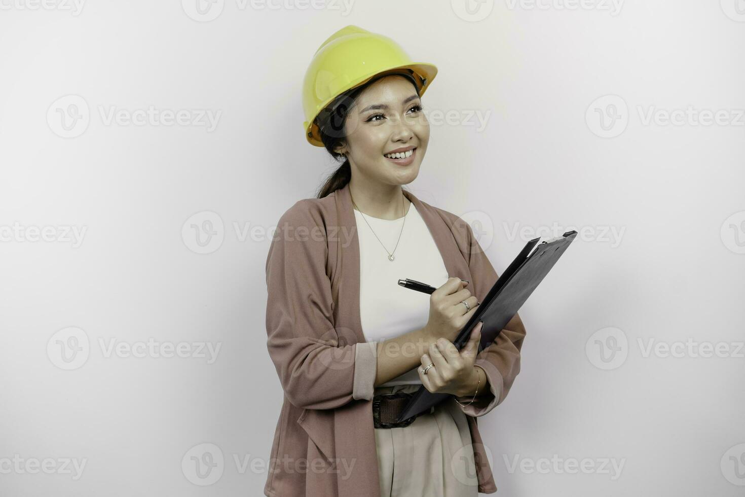 uma pensativo jovem ásia mulher empregado vestindo segurança capacete enquanto segurando uma prancheta e olhando a parte, de lado para cópia de espaço, isolado de branco fundo. foto
